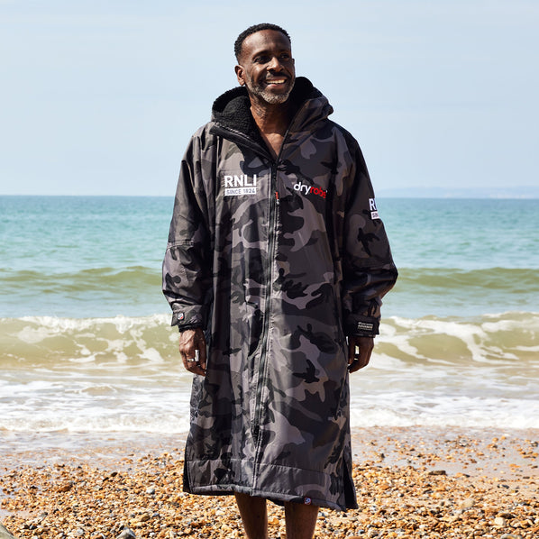 A man walking on the shoreline of a beach wearing a grey and black camouflage print dryrobe with the RNLI logo on the front.