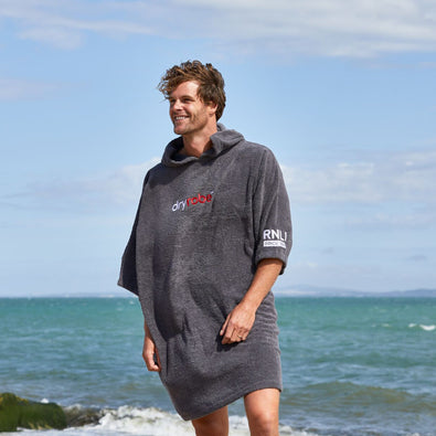 A man on a beach with the sea behind him wearing a grey RNLI dryrobe® Organic Towel Robe.