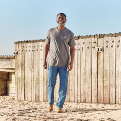A person wearing a charcoal t-shirt with an embroidered flag on the chest, standing in front of a wooden structure.