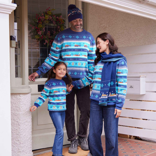 A family of three wearing matching RNLI blue and orange Christmas jumpers and accessories stands on a front porch.