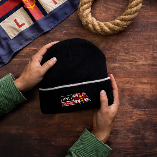 A pair of hands holding a navy beanie hat, with the RNLI Membership logo embroidered on  it. 