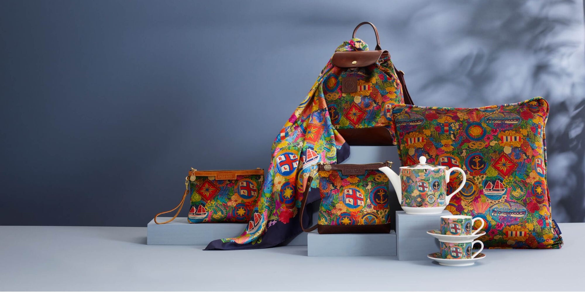 A selction of products from RNLI 200  birthday limited edition range, including stylish backpack, a cushion, a crossbody bag, a clutch bag, a silk scarf and a tea pot with two tea cups. Colourful print Designed by Emma Mawston.