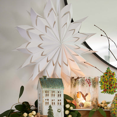 Decorative paper snowflake hanging above a small house and other festive items on a light background