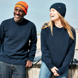 A man and a woman laughing together on the beach. They are both wearing the RNLI + Finisterre Polperro Jumper in Navy and matching beanies