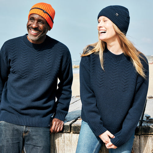A man and a woman laughing together on the beach. They are both wearing the RNLI + Finisterre Polperro Jumper in Navy and matching beanies