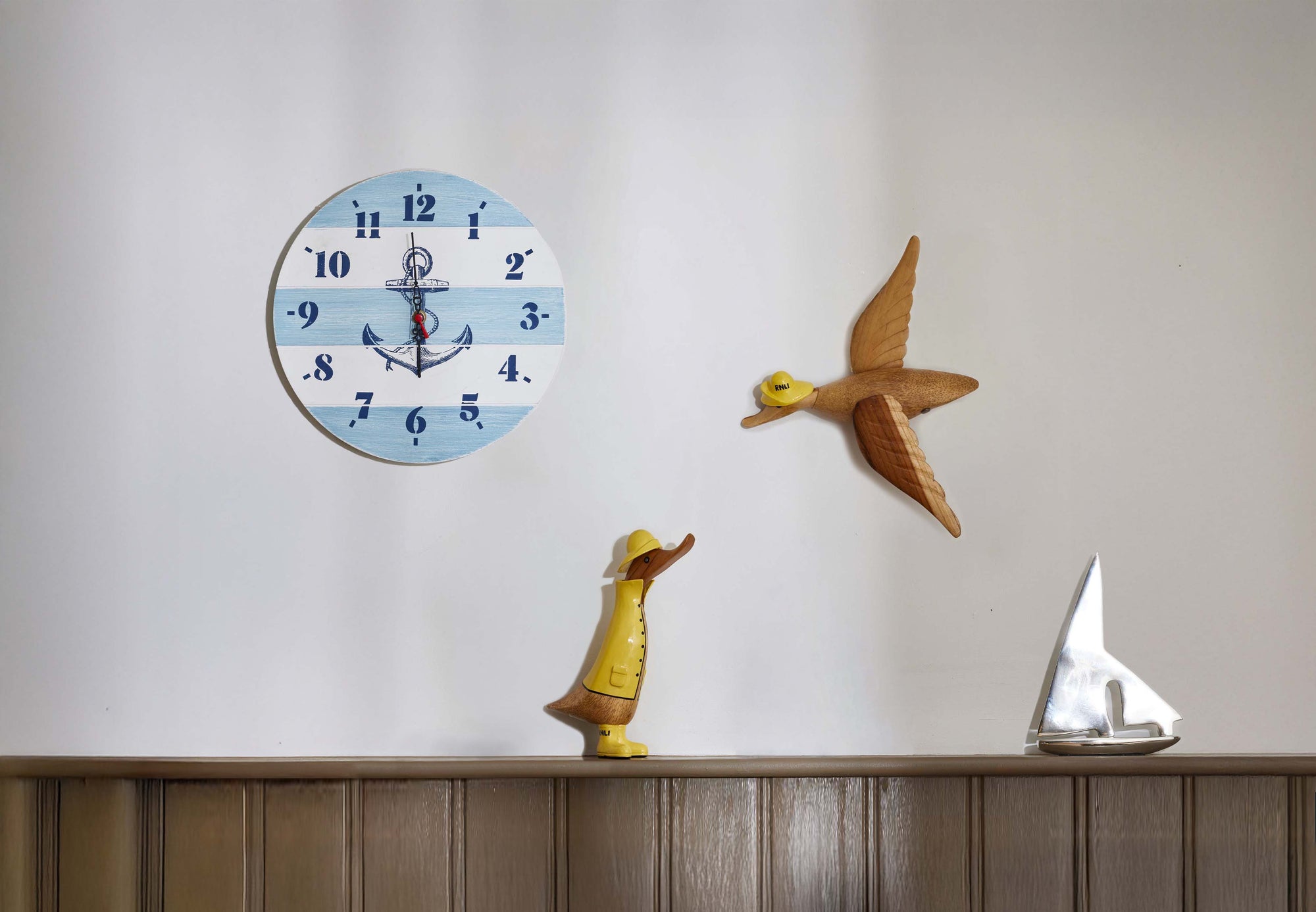 Decorative wall clock with nautical design on a wall, an RNLI George Duck in Sou'wester, DCUK RNLI Henry Flying Duck and Recycled Aluminium Yacht Sculpture.
