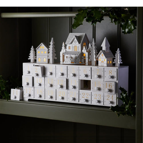A white, wooden advent with 24 boxes and a model house and trees on the top. 