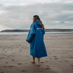 A woman walking on a stormy beach wearing a light blue dryrobe with long sleeves and a hood. 