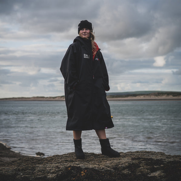 A woman standing on a rock with the sea behind, wearing a black dryrobe and a beanie hat. 