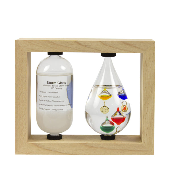 A Galileo thermometer and a storm bottle displayed in a wooden frame. The thermometer has colored bubbles inside a clear glass tube, and the storm bottle contains a mixture of liquids with colorful glass decorations representing weather indicators.
