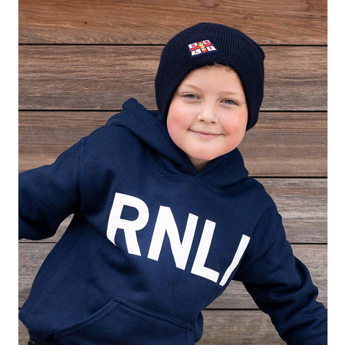 A young boy wearing a navy RNLI beanie and hoodie standing in front of a wooden-slatted wall. 