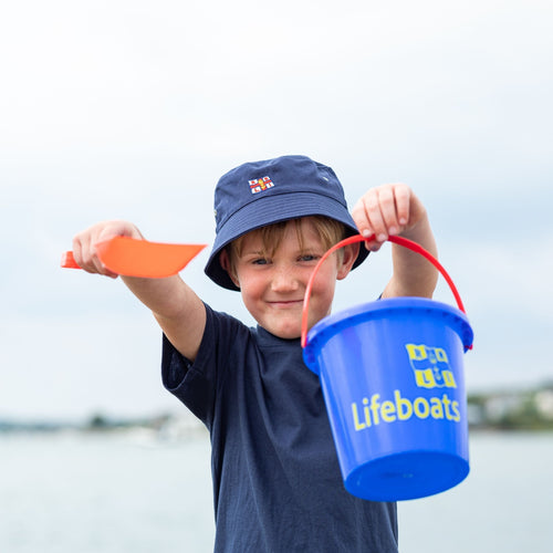 A child holding a bucket and spade at the beach and wearing a navy t-shirt with the RNLI flag.