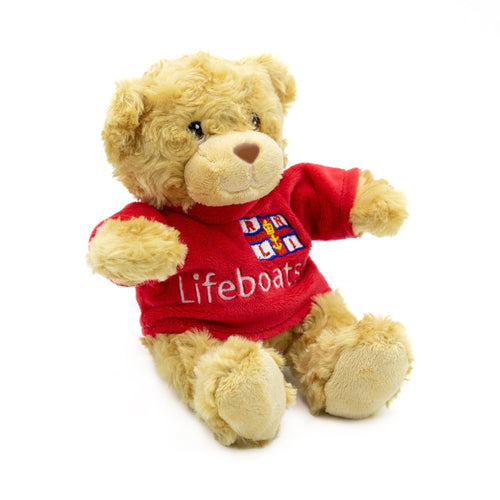 A light brown teddy bear wearing a bright red t-shirt with the RNLI Lifeboats logo on the front.