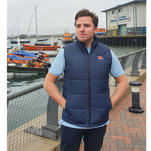 Man wearing the navy RNLI bodywarmer zipped up. He has his hands inside the two front pockets. In the background are RNLI lifeboats.