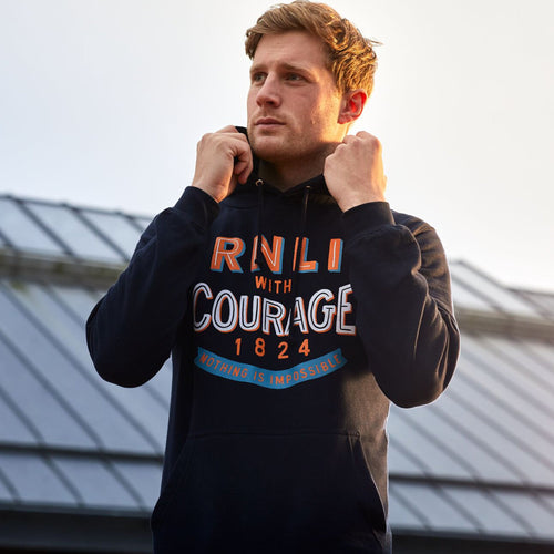 A man wearing a navy RNLI hoodie and pulling up the collar in the wind. The front of the hoodie has a print that reads, "RNLI with courage. 1824"