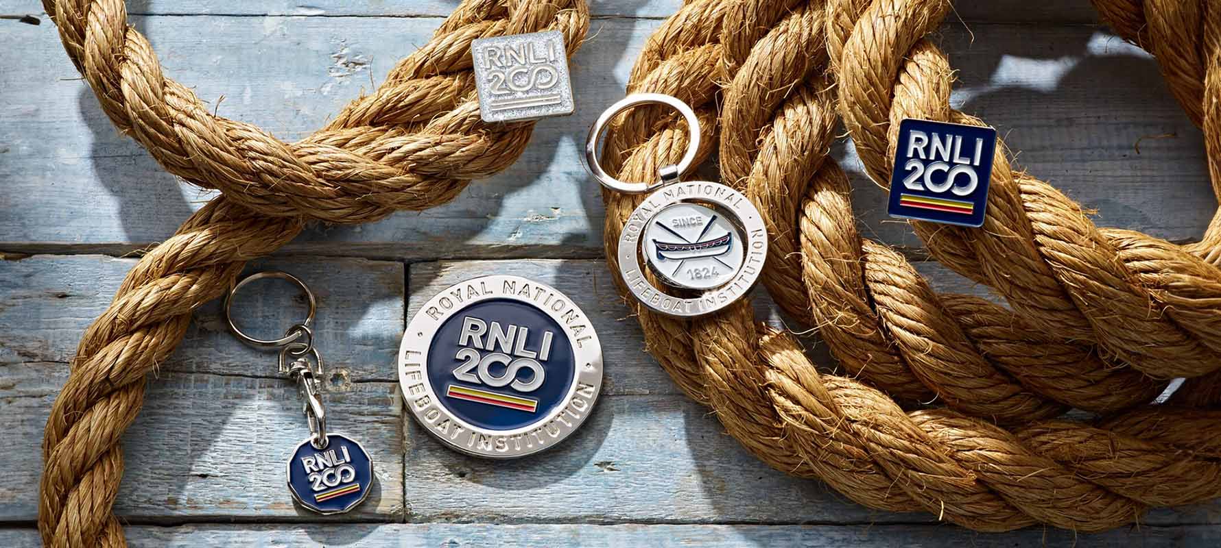 Overhead shot of keyrings and pin badges in the RNLI 200 range, laying over a rope on a blue wooden backdrop.