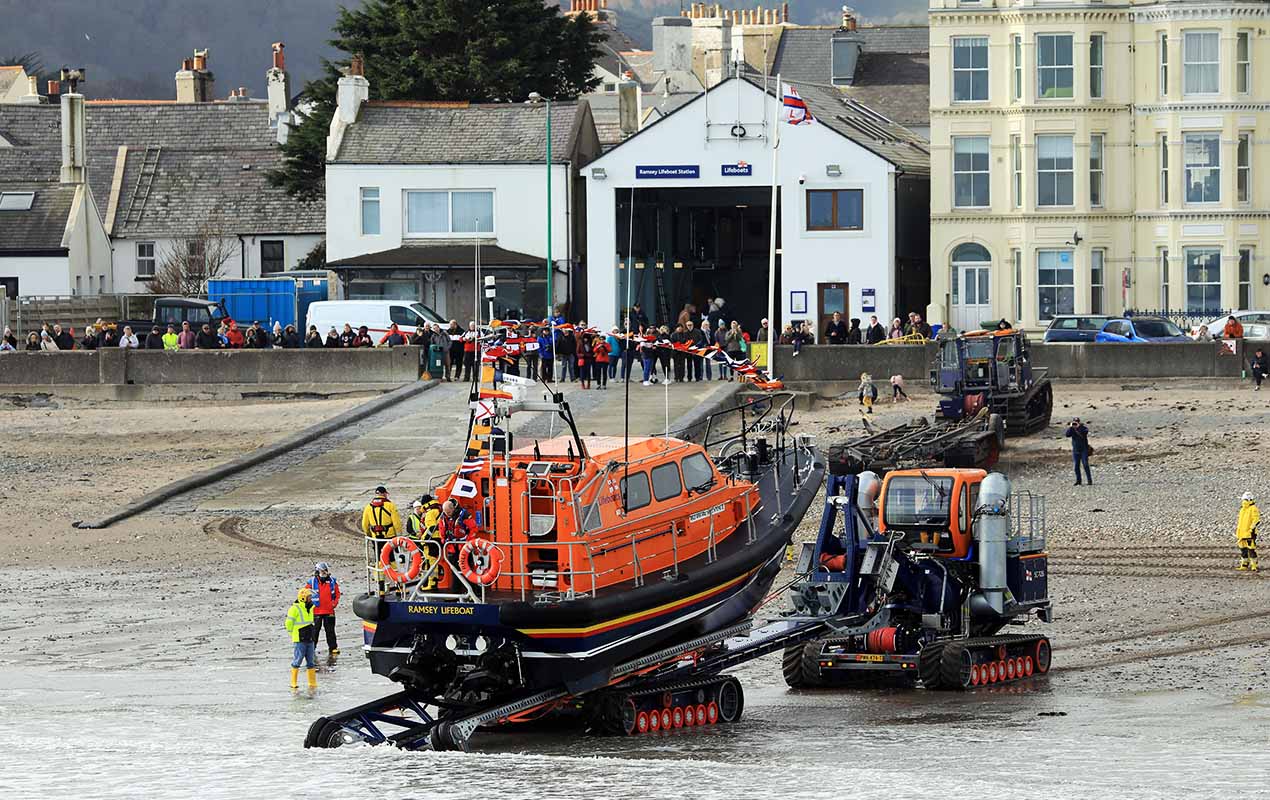 A Shannon class lifeboat is being recovered from the sea by a Shannon Launch and Recovery System, with Ramsey Lifeboat Station and crowds of people watching in the background.